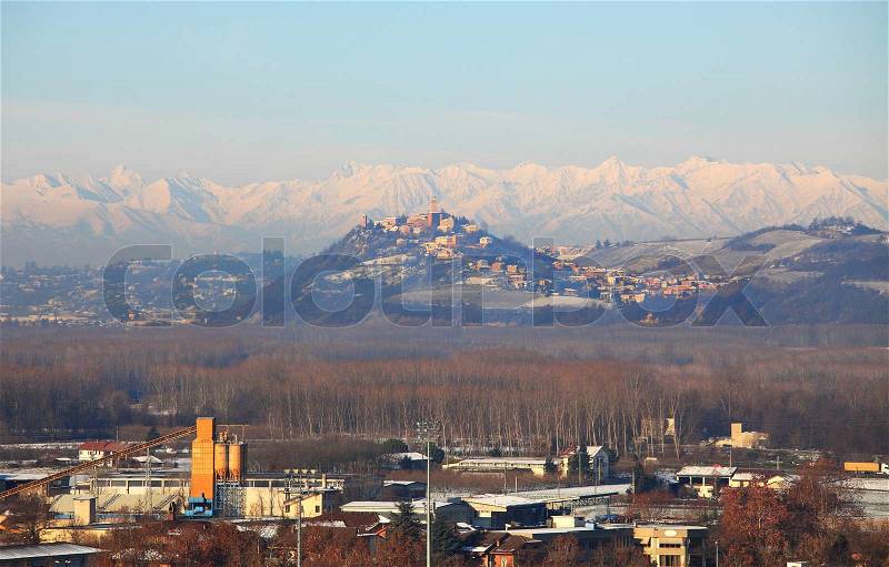 View on industrial zone and small town on snowy hill in front of snowy mountains in Piedmont, northern Italy, stock photo