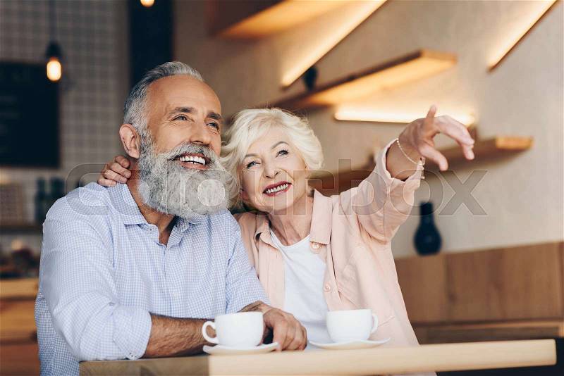 Smiling senior woman pointing away while drinking coffee together with husband in cafe, stock photo