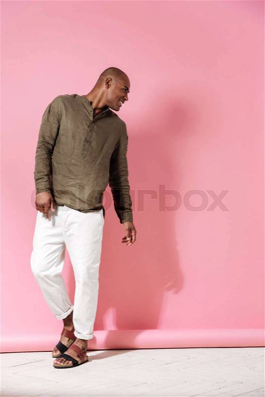 Full length image of happy african man posing in studio and looking away over pink background, stock photo