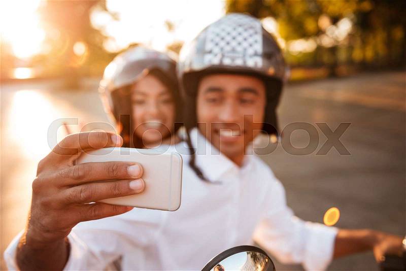 Happy african couple rides on modern motorbike outdoors and making selfie on smartphone. Focus on phone, stock photo