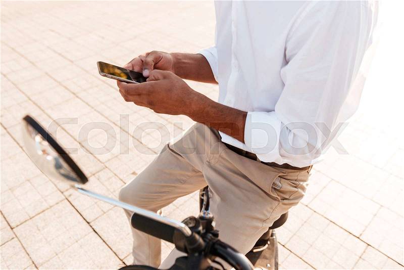 Cropped image of young african man sitting on modern motorbike outdoors and using smartphone, stock photo