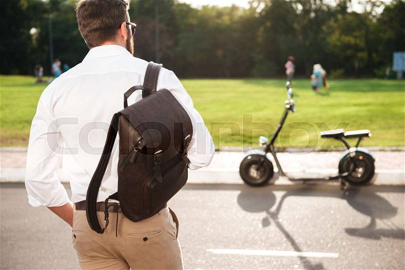 Back view of young man with backpack posing near the modern motorbike outdoors, stock photo