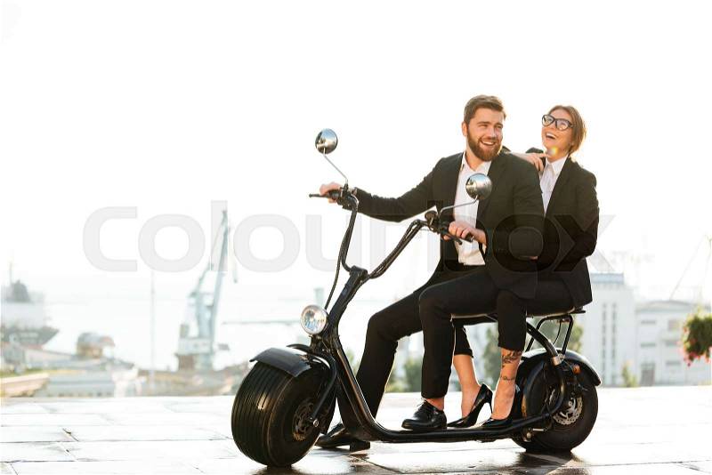 Full length side view image of laughing business couple sitting on modern motorbike in park and looking at the camera, stock photo