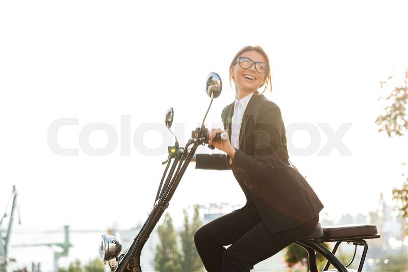 Side view of laughing business woman sitting on modern motorbike outdoors and looking away, stock photo