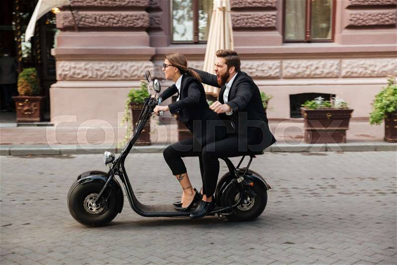 Cheerful excited couple wearing smart clothes riding motor bicycle on a city street, stock photo