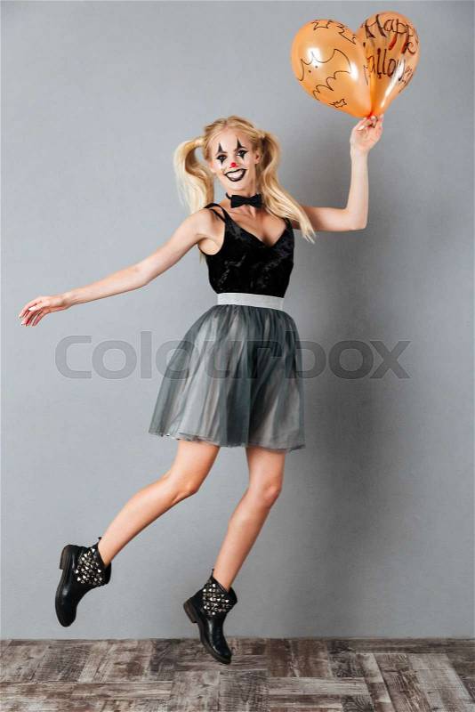 Full length image of smiling blonde woman in halloween make up jumping with balloons and looking at the camera over gray background, stock photo