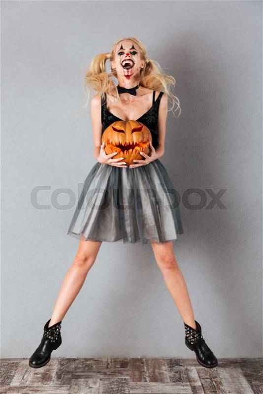 Full length portrait of a laughing crazy blonde woman in halloween clown make-up and blood streaks holding curved pumpkin and jumping isolated over gray background, stock photo