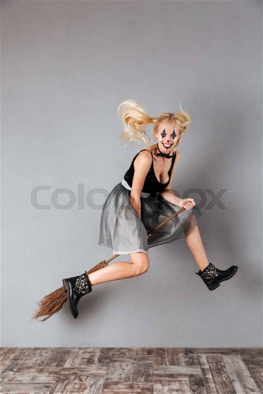 Full length portrait of a laughing crazy blonde woman in halloween clown make-up and blood streaks riding a broom while jumping isolated over gray background, stock photo