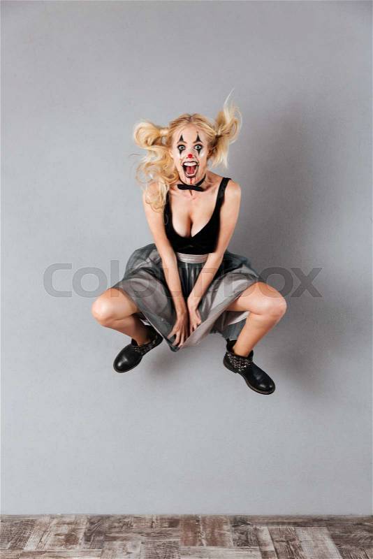 Full length portrait of a happy crazy blonde woman in halloween clown make-up and blood streaks jumping isolated over gray background, stock photo