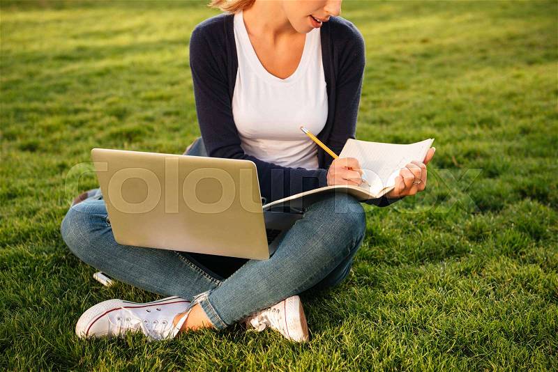 Cropped image of a girl student preparing for exams with textbook and laptop computer while sitting on the grass at park, stock photo