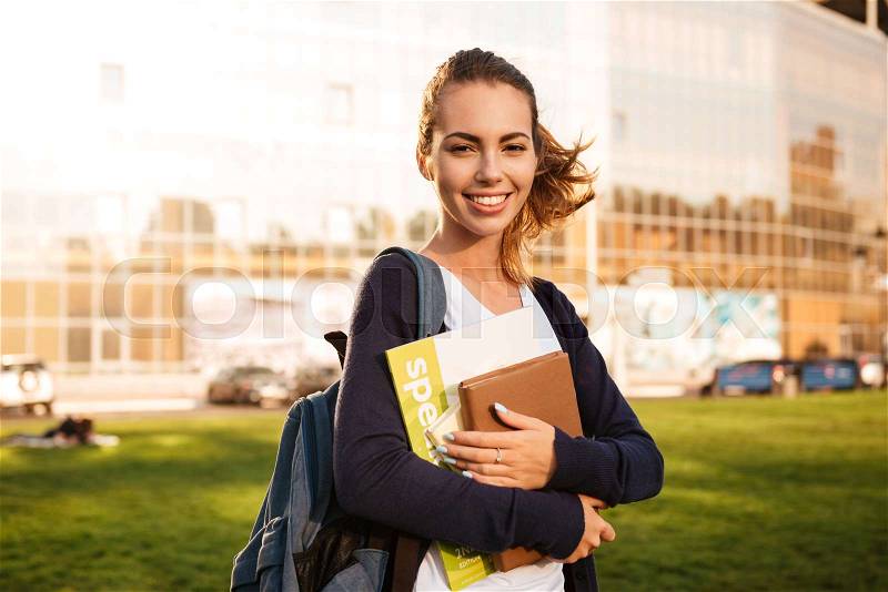 Portrait of a joyful brown-haired student girl standing with backpack and books and looking at camera outdoors, stock photo