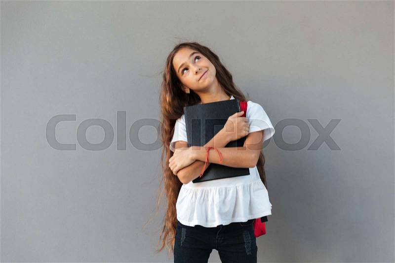Smiling pensive brunette schoolgirl with long hair hugging book and looking up over gray background, stock photo