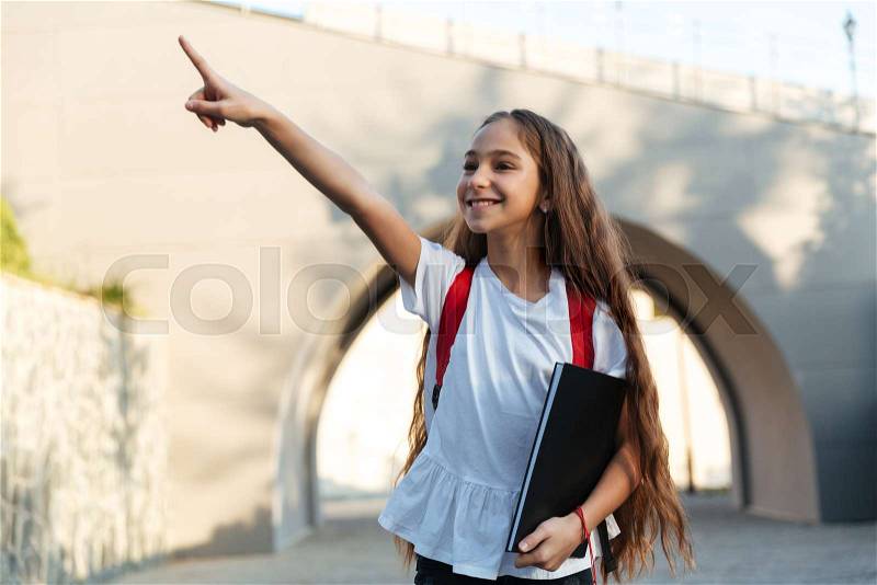 Smiling brunette schoolgirl posing outdoors pointing and looking away while holding book in hand, stock photo