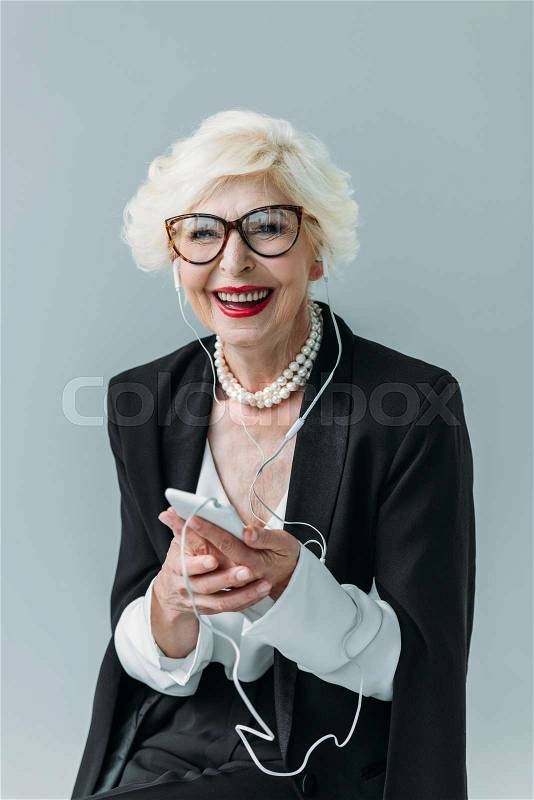 Smiling senior lady with pearl necklace listening music with earphones and smartphone, isolated on grey, stock photo
