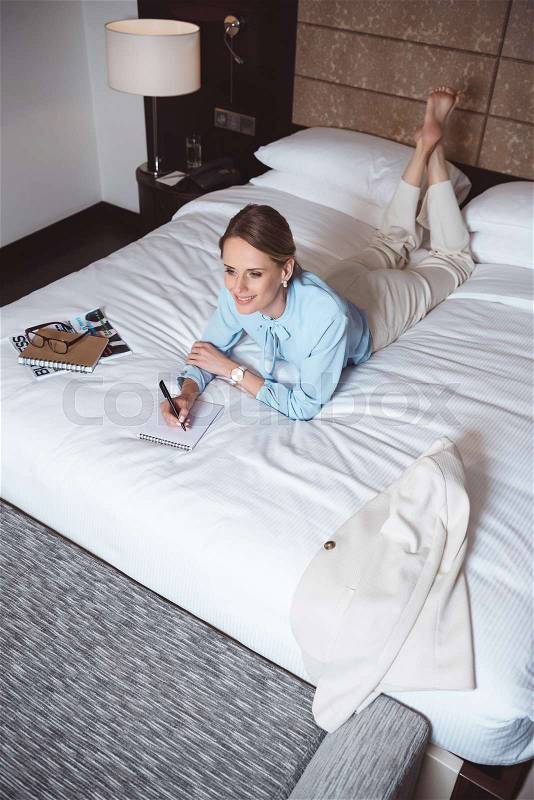 High angle view of smiling young businesswoman taking notes while lying on bed in hotel room , stock photo