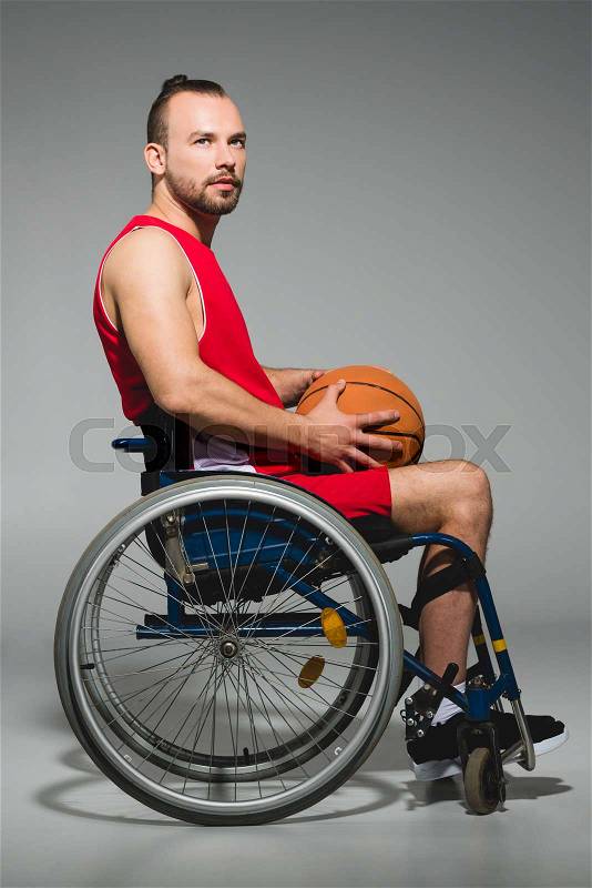 Young sportsman with basketball ball sitting in wheelchair and looking away, stock photo