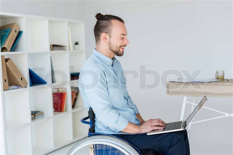 Smiling disabled businessman in wheelchair working with laptop in office, stock photo