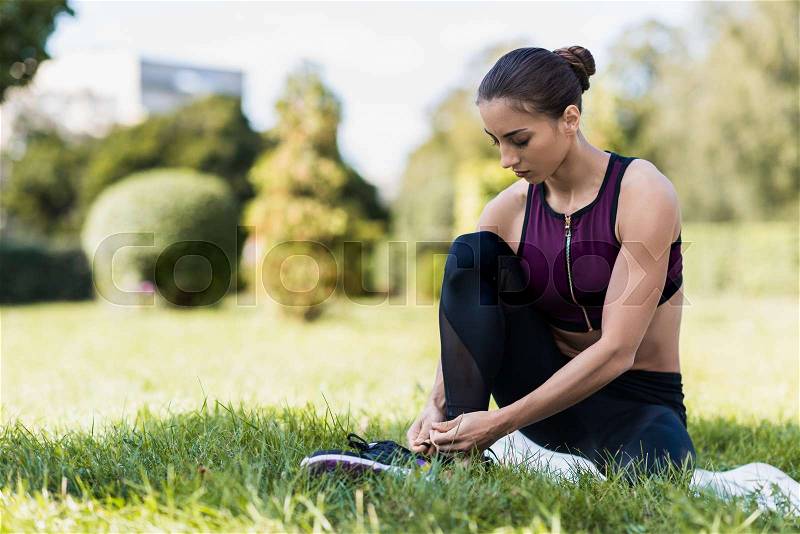 Young athletic woman lacing shoes while sitting on grass, stock photo