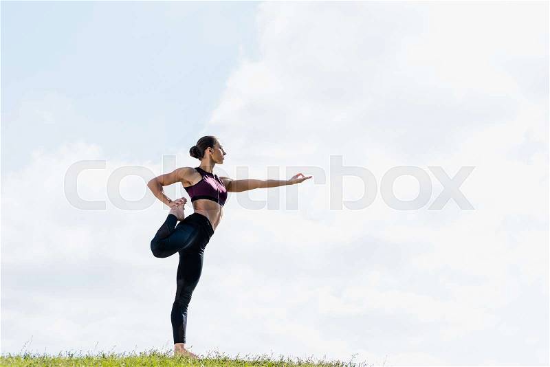 Young woman standing on one leg while practicing yoga outdoors on green grass, stock photo