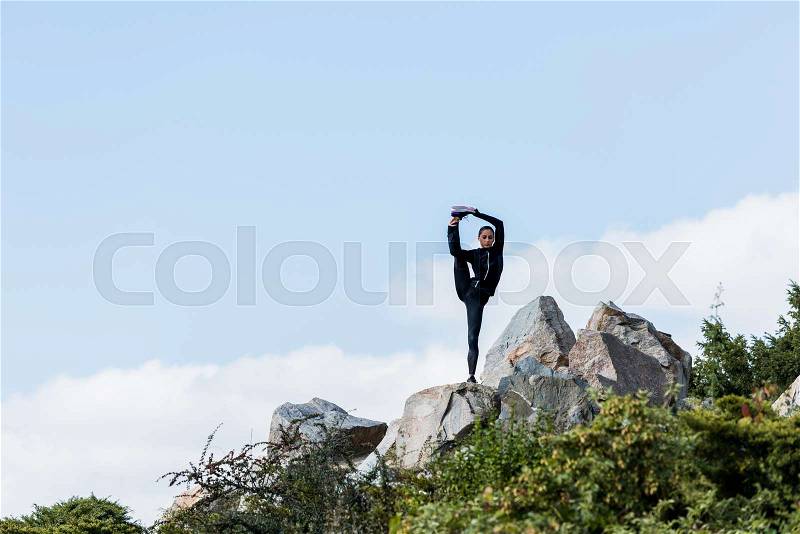 Young woman standing on one leg with other raised practicing yoga outdoors on rocks, stock photo