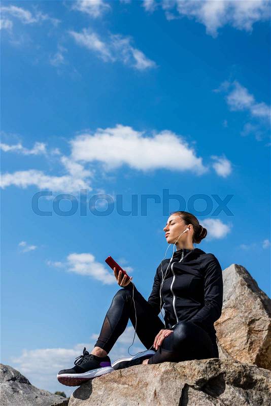 Young relaxed woman sitting on rocks and listening music in front of blue sky, stock photo
