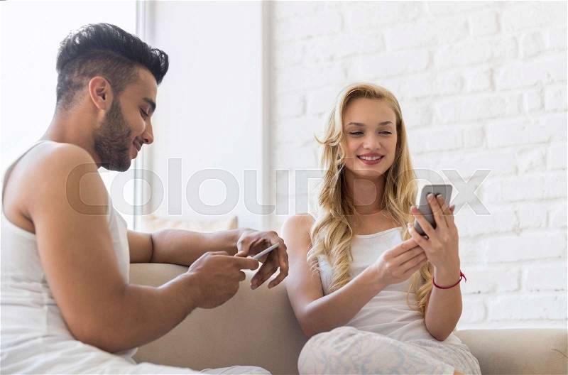 Young Beautiful Couple Sit On Coach Near Big Window, Using Cell Smart Phone Happy Smile Hispanic Man Woman Lovers, stock photo
