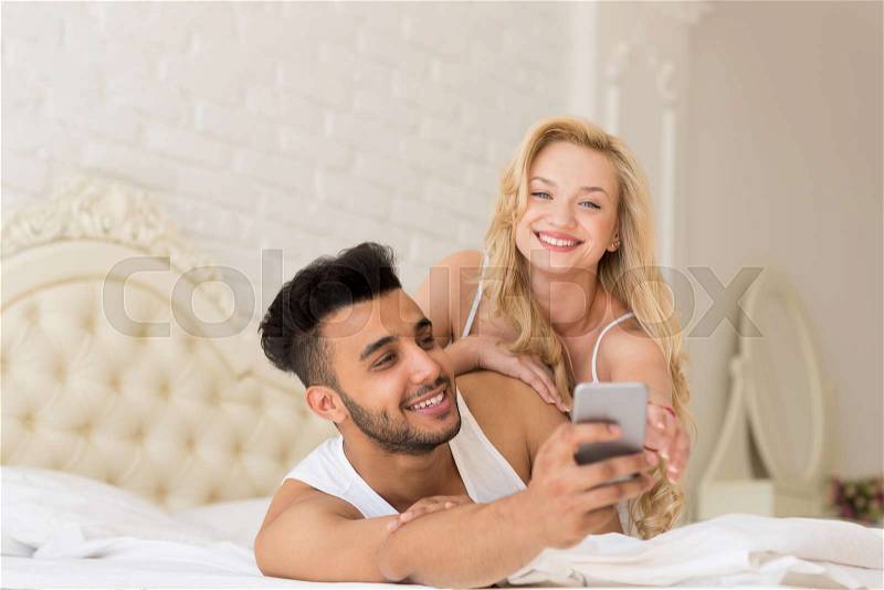Young Couple Lying In Bed, Happy Smile Hispanic Man Using Cell Smart Phone, Woman Trying To Take Away, Lovers Bedroom, stock photo
