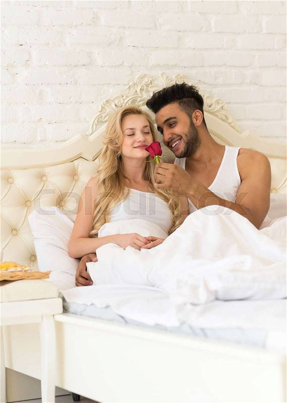 Young Couple Lying In Bed Eat Breakfast Morning With Red Rose Flower, Happy Smile Hispanic Man And Woman Lovers In Bedroom, stock photo