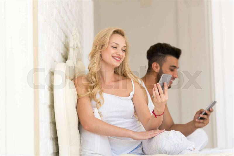Young Couple Sit In Bed, Happy Smile Hispanic Man And Woman Using Cell Smart Phone, Lovers In Bedroom, stock photo