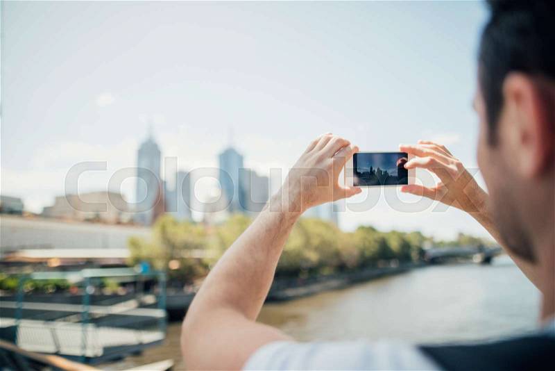 Millennial tourist is using his smart phone to take photos of the cityscape from a bridge in Melbourne, Victoria, stock photo