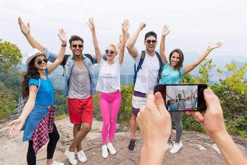 Cell Smart Phone Taking Photo Of Cheerful Tourist Group With Backpack Over Landscape From Mountain Top, People Posing With Raised Hands, stock photo