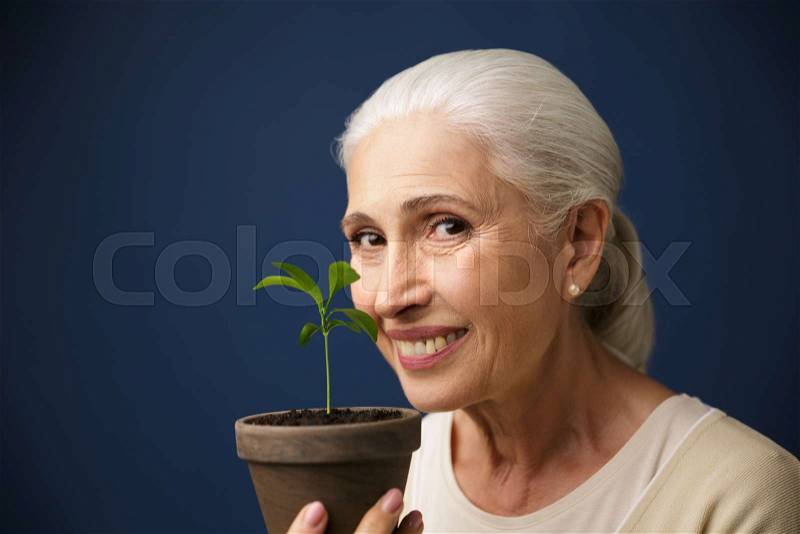 Close-up photo of happy aged woman showing young plant in the spot, looking at camera, over dark blue background, stock photo