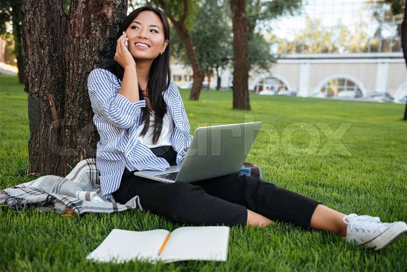 Young smiling chinese student speaking mobile phone, holding computer, sitting on grass, outdoor, stock photo