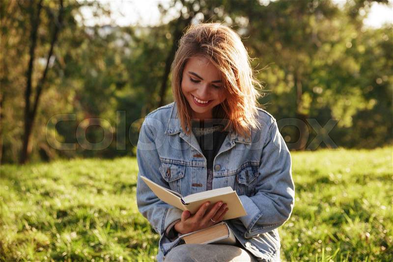 Cheerful young woman in jeans jacket reading a book in sunny park, stock photo