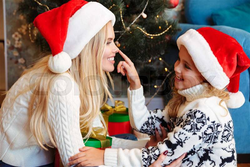 Cute little daughter in Santa\'s hat touching nose of her smiling mother at Christmas tree, stock photo