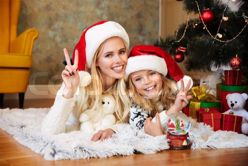 Young smiling mother with her daughter in Santa's hat looking at camera, showing peace gesture while lying on the floor near Christmas tree, stock photo