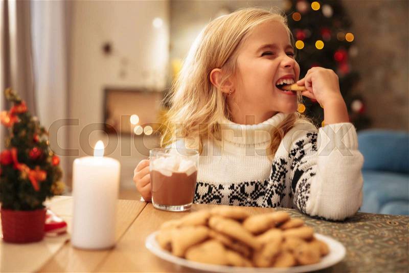 Cute little girl drinking cacao and eating cookie at home, stock photo