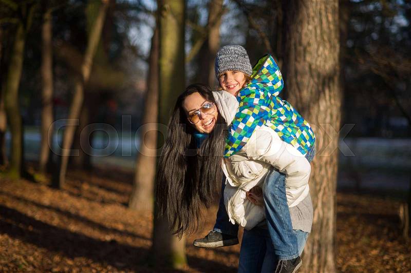 Young woman bears on a back of the boy of 9-10 years. Mum and the son on walk in autumn park. Serene autumn day, good mood, stock photo