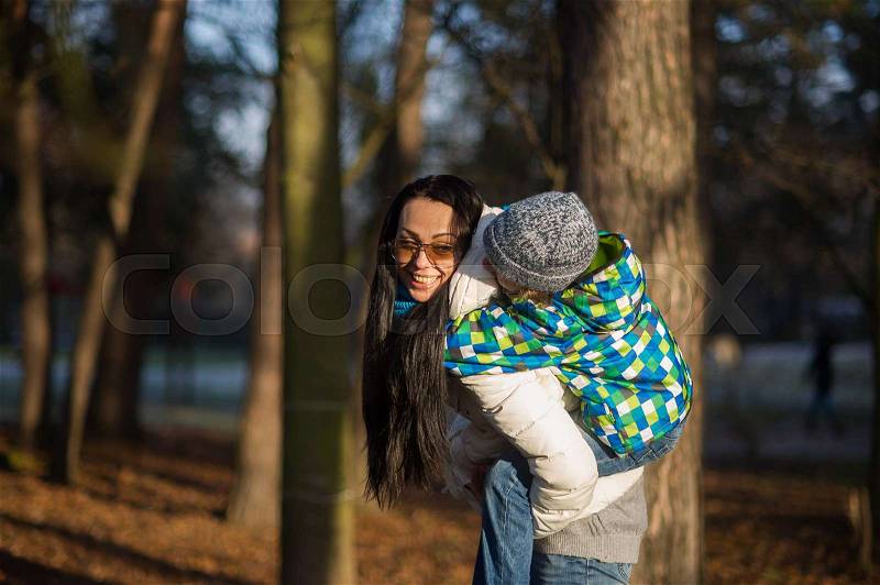 Young woman bears on a back of the boy of 9-10 years. Mum and the son on walk in autumn park. Serene autumn day, good mood, stock photo
