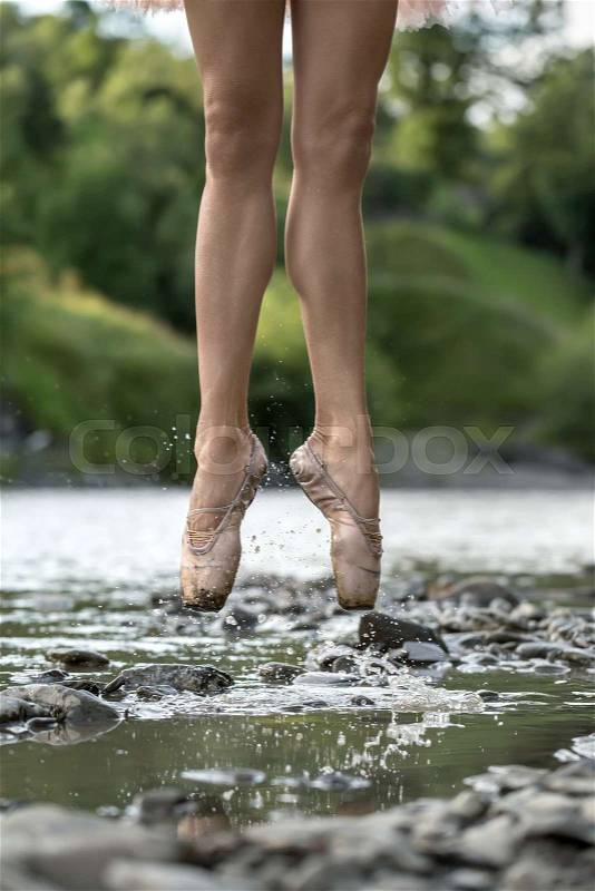 Incredible ballerina jumping in the shallow river on the background of the green shore. She wears beige pointes. Water splashes spreading around her legs. Closeup. Vertical, stock photo