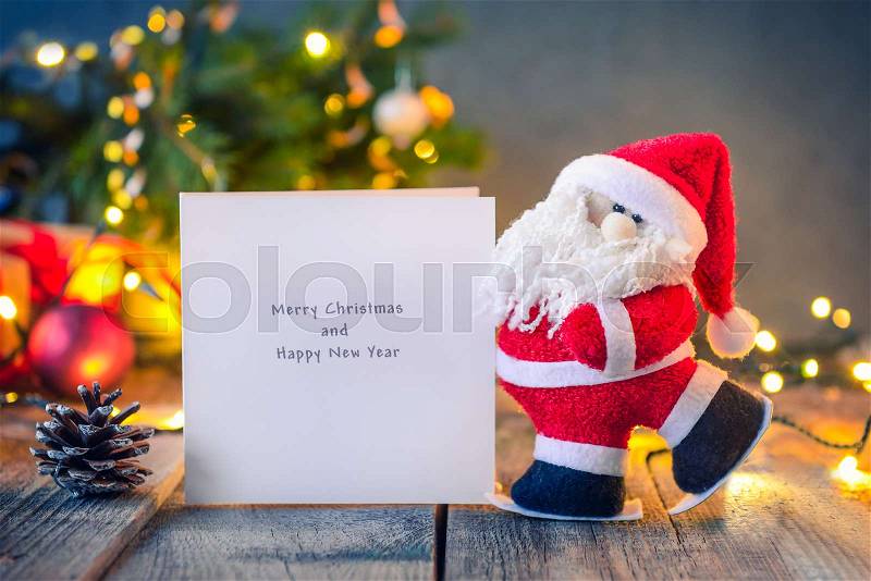 Decorative Santa Claus carrying greeting card with best wishes on the festive dark background with Christmas tree, lights and decor on the old wood. New year vintage greeting card. Selective focus, stock photo