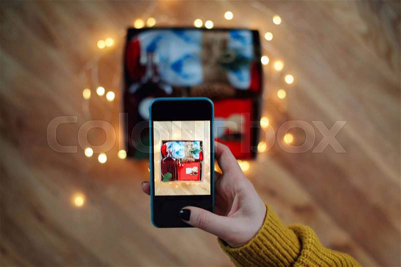 Woman taking photo of Christmas gift box with smartphone. Instagram photography blogging workshop concept. A girl hanging a phone taking a photo of present on wooden table, stock photo