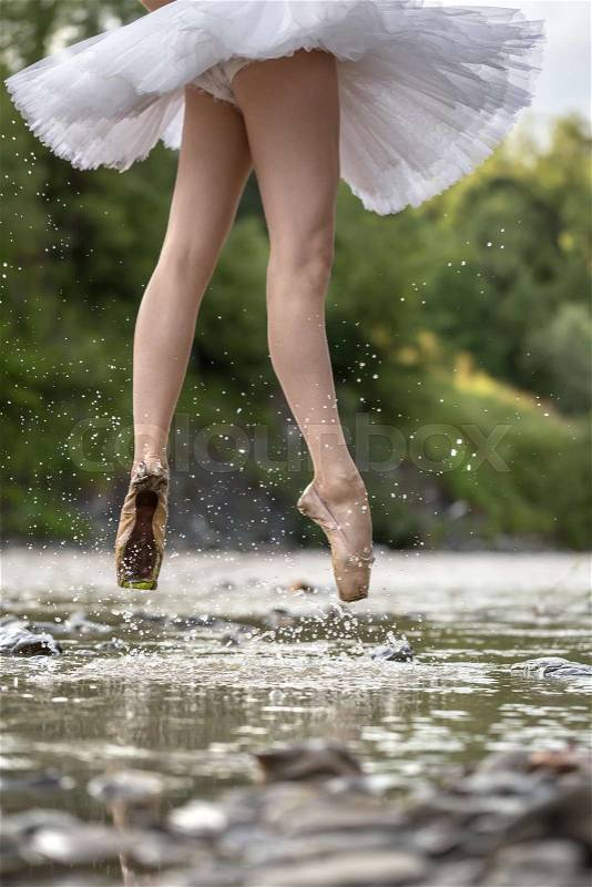 Sensual ballerina jumping in the shallow river on the background of the green shore. She wears a white tutu and beige pointes. Water splashes spreading around her legs. Closeup. Vertical, stock photo