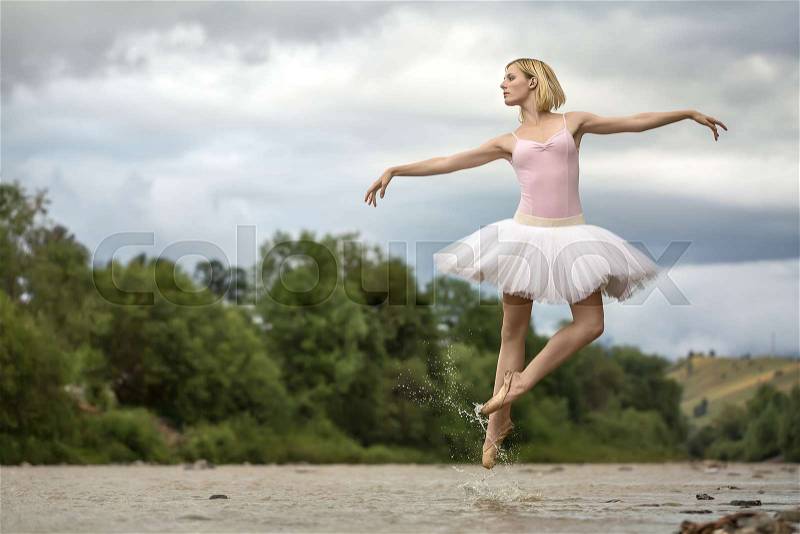 Delightful ballerina jumping in the shallow river on the background of green shore and cloudy sky. She wears white tutu, pink leotard and beige pointes. Water splashes spreading around her legs, stock photo