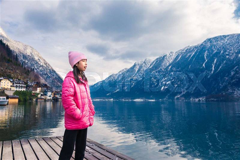 Girl in pink winter cloth staning by a lake with blurred town and mountain range across the water in background, room for text, good background for travel or family theme concept, stock photo