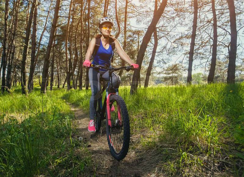 One young woman - an athlete in a helmet riding a mountain bike outside the city, on the road in a pine forest on a summer day, stock photo