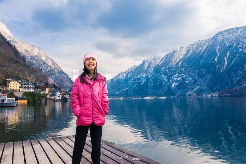 Happy asian girl in pink winter cloth staning by a lake with blurred town and mountain range across the water in background, room for text, good background for travel or family theme concept, stock photo
