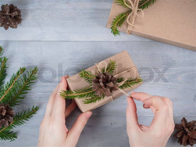 Female hands unpacking christmas holiday handmade present in craft paper with spruce branches on gray wooden table. Top view. Christmas background, stock photo