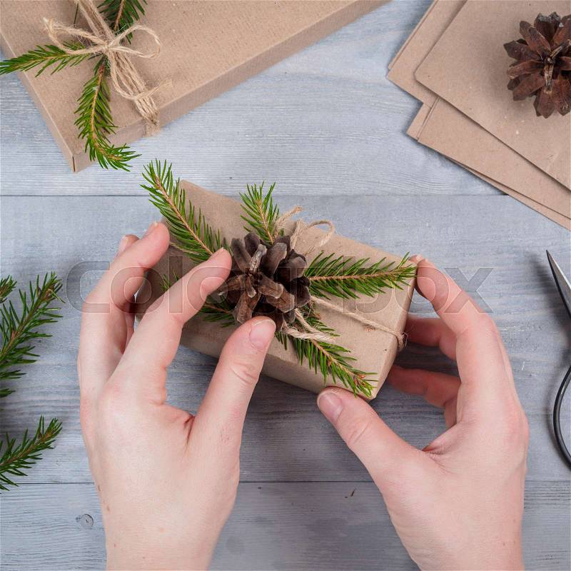 Female hands give wrapped christmas holiday handmade present in craft paper. Spruce branches and tools on gray wooden table. Top view.Workplace for preparing handmade decorations.Christmas background, stock photo