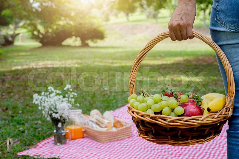 Picnic wicker basket with food, bread, fruit and orange juice on a red and white checked cloth in the field with green nature background. Picnic concept, stock photo